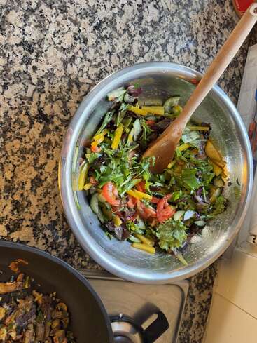 Ein frischer Gemüsesalat mit verschiedenen Blattsalaten, Tomaten, gelber Paprika und Gurken in einer Metallschüssel. Daneben liegen ein Holzlöffel und ein gekochtes Gericht.