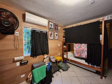 A cozy hostel room with wooden walls features a bunk bed, air conditioner, fan, window with curtains, travel bags, colorful towel, and framed artwork.