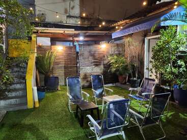 A cozy outdoor patio with string lights, five blue chairs, a small table, artificial grass, potted plants, wooden fence, and stairs on the left side.