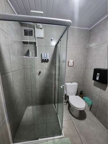 This is a modern bathroom with grey tiles, a glass-enclosed shower, dispensers, toilet, trash can, wall-mounted black box, and a green floor mat.