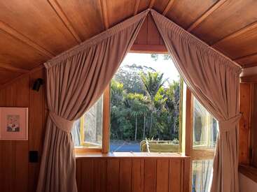 La luz del sol se cuela por una ventana triangular con cortinas beige abiertas en una acogedora cabaña de madera, dejando ver los frondosos árboles verdes y el cielo azul del exterior, creando tranquilidad.