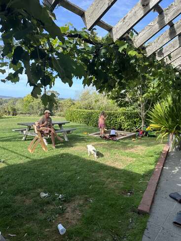Un hombre se relaja en un banco de picnic mientras los niños juegan en un arenero. Un perro pequeño explora el patio cubierto de hierba bajo una frondosa pérgola, disfrutando del sol.