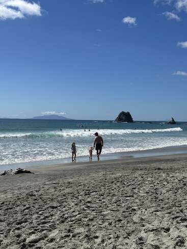 Un hombre y dos niños caminan por una playa de arena hacia el océano, con cielos azules, suaves olas, nadadores lejanos e islas rocosas al fondo.