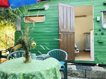 A cozy green caravan with an open wooden door sits beside a table covered in a floral cloth, vase of wildflowers, chairs, books, and a mug.