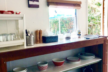 This image depicts a kitchen shelf with various glassware, bowls, plates, utensils, and condiments. A window above reveals greenery outside, creating a bright, inviting atmosphere.