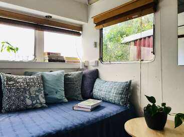 A cozy nook with patterned pillows, books on the windowsill, indoor plants, large windows with wooden blinds, and a blue-covered bed create a serene reading corner.