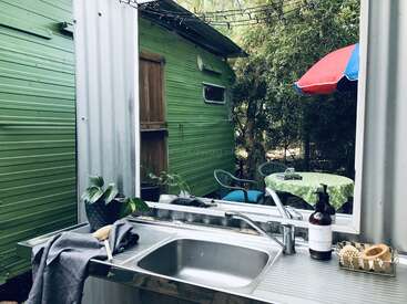 An outdoor sink with a mirror overlooks a green trailer, garden table, and chairs. There’s a colorful umbrella, soap dispenser, and plants, creating a cozy atmosphere.