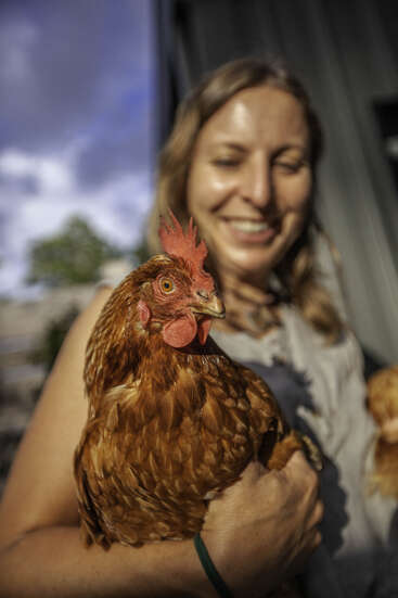 A smiling woman holds a brown chicken in her arms. The chicken is in focus, while the woman stands in soft, cheerful sunlight, slightly blurred.