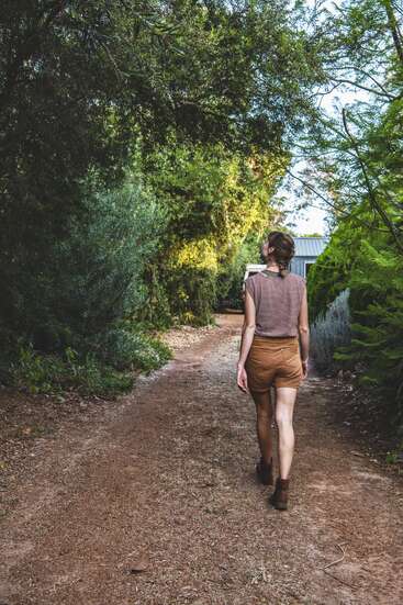 A woman in brown shorts and a striped shirt walks along a dirt path surrounded by lush green trees and foliage, heading towards a small building.