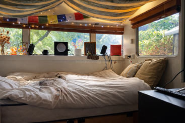 Un acogedor dormitorio con una cama grande, almohadas de colores, cálidas claraboyas, banderas de oración y una decoración vibrante. Las ventanas dejan ver la exuberante vegetación del exterior, creando un ambiente tranquilo.