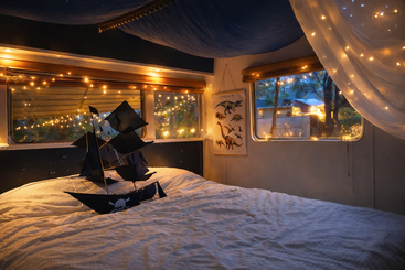 Un acogedor dormitorio con una decoración de barco pirata sobre la cama, iluminado por cálidas luces de cuerda y telas drapeadas, con póster de dinosaurio y resplandor nocturno exterior.