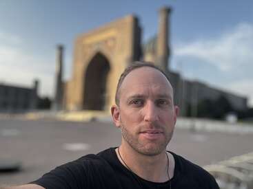 A man takes a selfie outdoors with a large, ornate, ancient building in the background. The structure features tall towers and an arched entrance. Sky is clear.