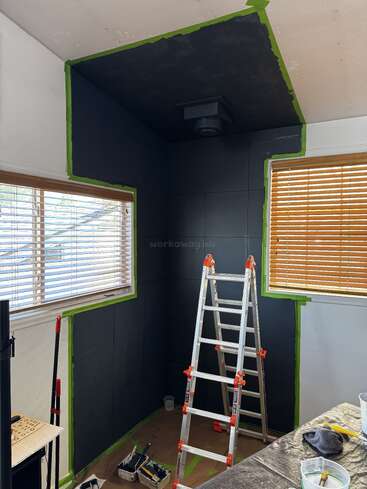 A room under renovation, black walls and ceiling partially painted, outlined with green painter’s tape. A ladder, painting tools, and two windows with blinds visible.