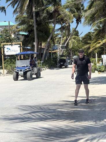A man in black stands on a sunny, tropical street lined with palm trees. A golf cart drives by, and colorful buildings are visible behind him.
