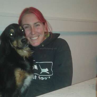 The image shows a woman with red hair and a black hoodie, sitting at a table with a dog, smiling at the camera.