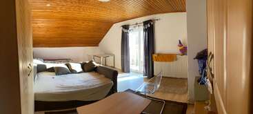 Um quarto aconchegante no sótão com teto de madeira, cama grande, sofá, mesa, cadeira de vidro, guarda-roupa e portas da varanda com cortinas escuras que deixam a luz do sol entrar.