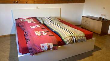 Esta imagem mostra um quarto moderno com uma cama branca, dois edredons coloridos que não combinam entre si, lençóis vermelhos e uma cômoda de madeira contra uma parede de cor clara.