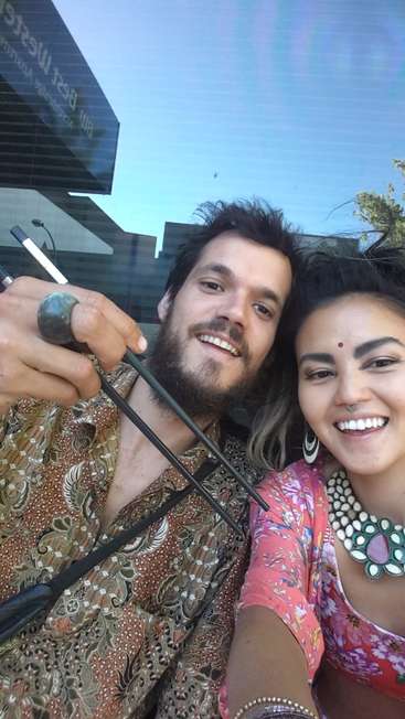 A imagem mostra um homem e uma mulher posando para uma selfie, com o homem segurando um cachimbo e usando uma camisa estampada, e a mulher usando uma blusa floral rosa.