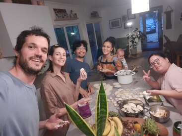 Um grupo de amigos faz bolinhos de massa juntos em uma cozinha aconchegante. Há um bebê, muitos sorrisos, preparações de alimentos, plantas e uma atmosfera amigável e descontraída.