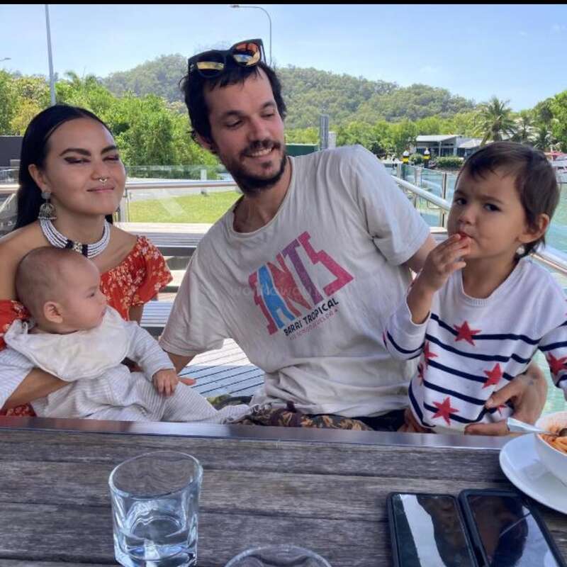 Uma família de quatro pessoas senta-se ao ar livre em uma mesa perto da água, aproveitando um dia ensolarado. Os pais sorriem enquanto as crianças comem e relaxam juntas.