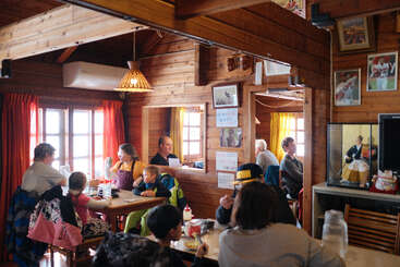 Un acogedor restaurante con aspecto de cabaña de madera, lleno de familias y amigos que disfrutan de la comida. Una iluminación cálida, cortinas de colores, cuadros enmarcados y un ambiente acogedor y hogareño completan el cuadro.