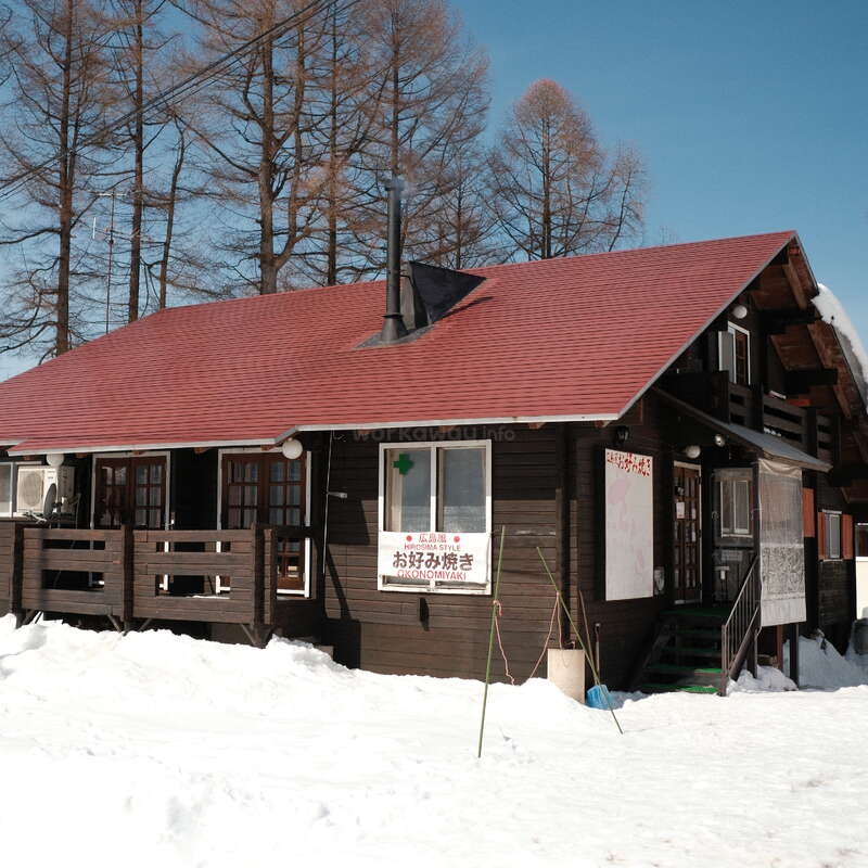 Una acogedora cabaña de madera con techo rojo se alza en medio de un terreno nevado, rodeada de altos árboles sin hojas. Un cartel anuncia okonomiyaki al estilo de Hiroshima. Sobre ella, un cielo azul y despejado.