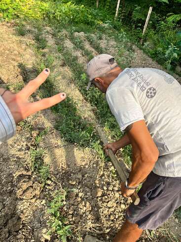 A man is working in a garden, tending to plants with a tool, while another person shows a peace sign with painted nails in the foreground.