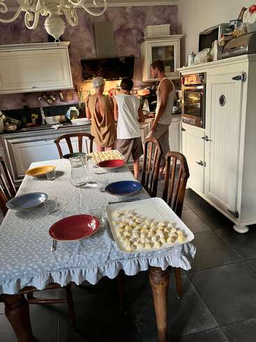 Three people prepare food together in a cozy kitchen. A table set for four with colorful plates and homemade gnocchi suggests a warm, communal meal ahead.