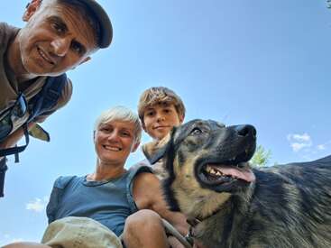 Three people and a happy dog pose for a selfie outdoors. The sky is blue. Everyone looks cheerful, enjoying a sunny day and time together.