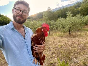A man with glasses and a beard, wearing a light blue shirt, holds a large red rooster outdoors in a sunlit, green rural landscape.