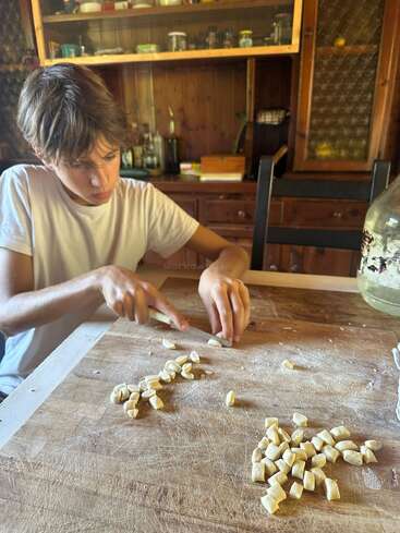 A boy in a white t-shirt is carefully cutting dough into small pieces on a wooden board in a rustic kitchen, making homemade gnocchi pasta.