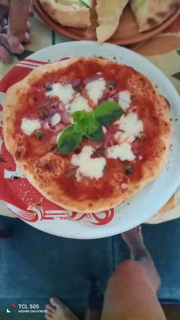 A freshly baked pizza with tomato sauce, mozzarella, red onion, capers, and basil sits on a white plate. Bare feet and hands are visible nearby.