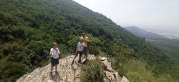 Three people stand on a rocky outcrop surrounded by lush green mountains. The sky is clear, and a distant landscape is visible beyond the hills.