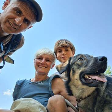 Drei Menschen und ein glücklicher Hund posieren für ein Selfie im Freien. Der Himmel ist blau. Alle sehen fröhlich aus, genießen den sonnigen Tag und die gemeinsame Zeit.