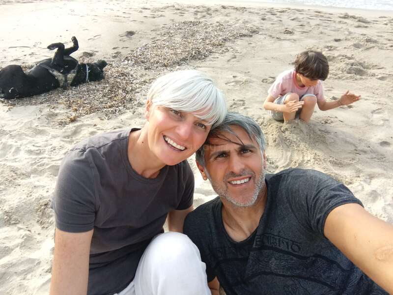 The image shows a family of three on a sandy beach, with a dog lying in the background, and the man and woman posing for a selfie.