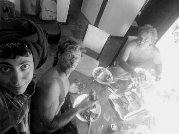 Trois personnes partagent un repas à une table. Deux d'entre elles sont torse nu, l'une porte un bandeau et sourit à la caméra. La nourriture, les boissons et les ustensiles sont visibles.