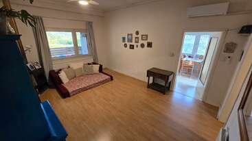 Uma sala de estar aconchegante com piso de madeira, um sofá marrom, arte na parede, armário azul, janela com cortinas, mesa pequena e uma área de jantar iluminada adjacente.