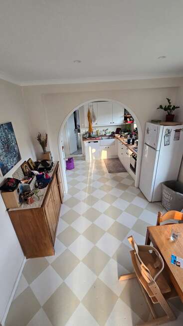 Esta imagem mostra uma cozinha aconchegante com piso xadrez, móveis de madeira, arco, plantas, uma geladeira, alguma desordem nas superfícies e luz do sol que atravessa o espaço.