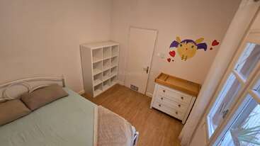 Cette chambre confortable comprend un lit simple avec des oreillers, une étagère blanche, une commode avec un matelas à langer, des œuvres d'art murales et une grande fenêtre lumineuse avec des rideaux.