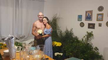 Un homme torse nu et une femme en robe bleue s'embrassent amoureusement à l'intérieur, entourés de fleurs, de plantes vertes, de verrerie et d'œuvres d'art sur le mur derrière eux.