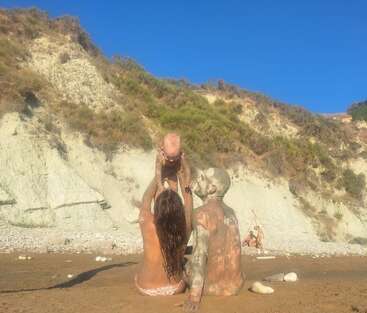 Une famille est assise sur une plage de sable, couverte de boue. La femme soulève un bébé en s'amusant, tandis que l'homme, lui aussi couvert de boue, regarde la scène d'un air heureux. Des collines s'élèvent derrière.