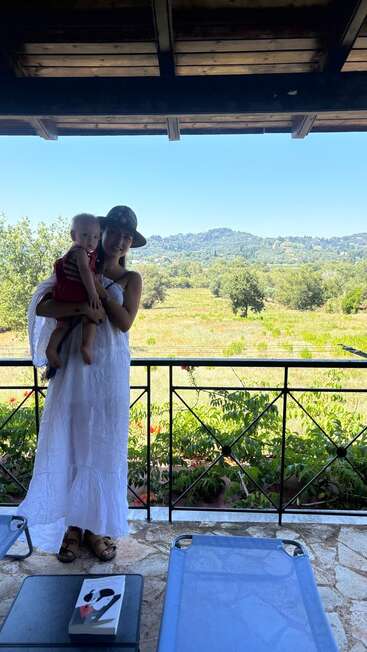 Une femme vêtue d'une robe et d'un chapeau blancs tient un enfant dans ses bras sur un patio en pierre. Derrière eux, des champs et des collines verdoyantes s'étendent sous un ciel bleu clair.