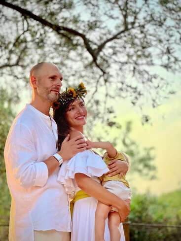 Une famille heureuse se tient à l'extérieur, éclairée par la lumière chaude du soleil. L'homme et la femme sont vêtus de blanc ; la femme porte une couronne de fleurs et tient affectueusement un petit enfant dans ses bras. Un moment paisible et joyeux.