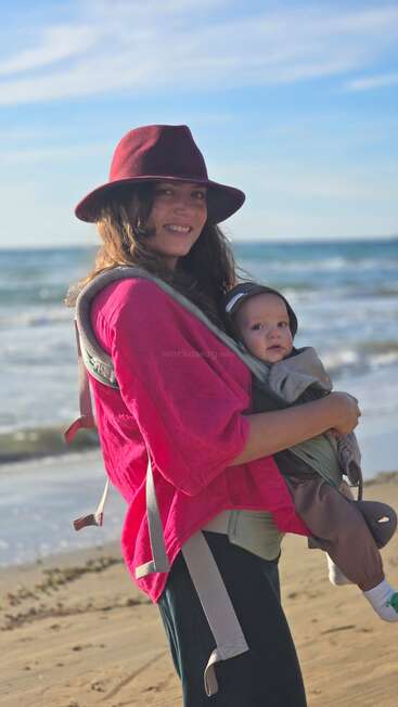 Une femme souriante, coiffée d'un chapeau rouge et vêtue d'une chemise rose, porte un bébé dans un porte-bébé avant tout en marchant sur une plage de sable près de l'océan.