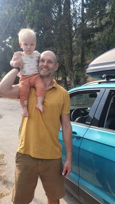 Un homme souriant en chemise moutarde tient un petit enfant sur son épaule, debout à côté d'une voiture bleue avec des arbres et de la verdure en arrière-plan.