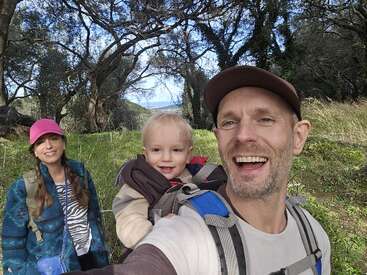 Une famille souriante profitant d'une randonnée dans la nature : le père porte un jeune enfant dans un sac à dos porte-bébé, la mère porte un chapeau rose, le tout au milieu des arbres et de la verdure.