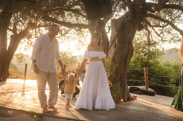 Une famille rayonnante marche pieds nus sur une terrasse en bois, à l'ombre de grands arbres, au coucher du soleil. La femme, vêtue d'une robe blanche et d'une couronne de fleurs, tient la main d'un petit enfant.