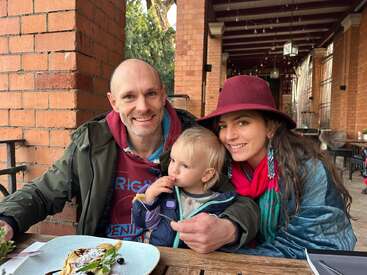Une joyeuse famille de trois personnes est assise à la terrasse d'un restaurant. Le père sourit, la mère porte un chapeau rouge et l'enfant mange avec curiosité.