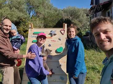 Quatre adultes, dont l'un tient un bébé dans ses bras et un autre un chiot, se tiennent debout en souriant devant un petit mur d'escalade en bois, à l'extérieur, par une journée ensoleillée.