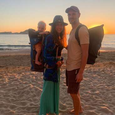 Uma família sorridente está em uma praia de areia ao pôr do sol. A mulher carrega um bebê em uma mochila porta-bebê, enquanto o homem tem uma mochila grande.